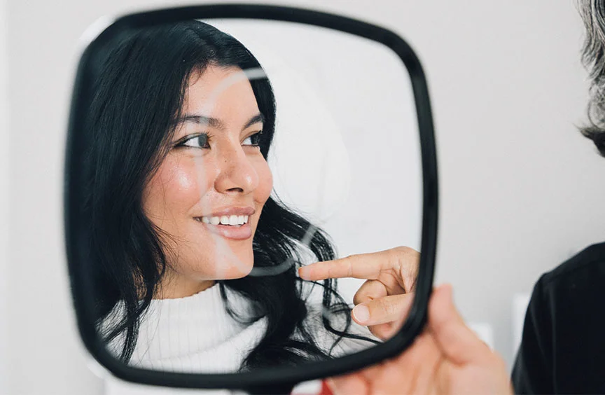A close-up shot of a woman with long black hair smiling into a handheld mirror as a doctor points at her face. She is wearing a white shirt, appearing engaged and content during the consultation. - Kybella in Bryn Mawr, PA