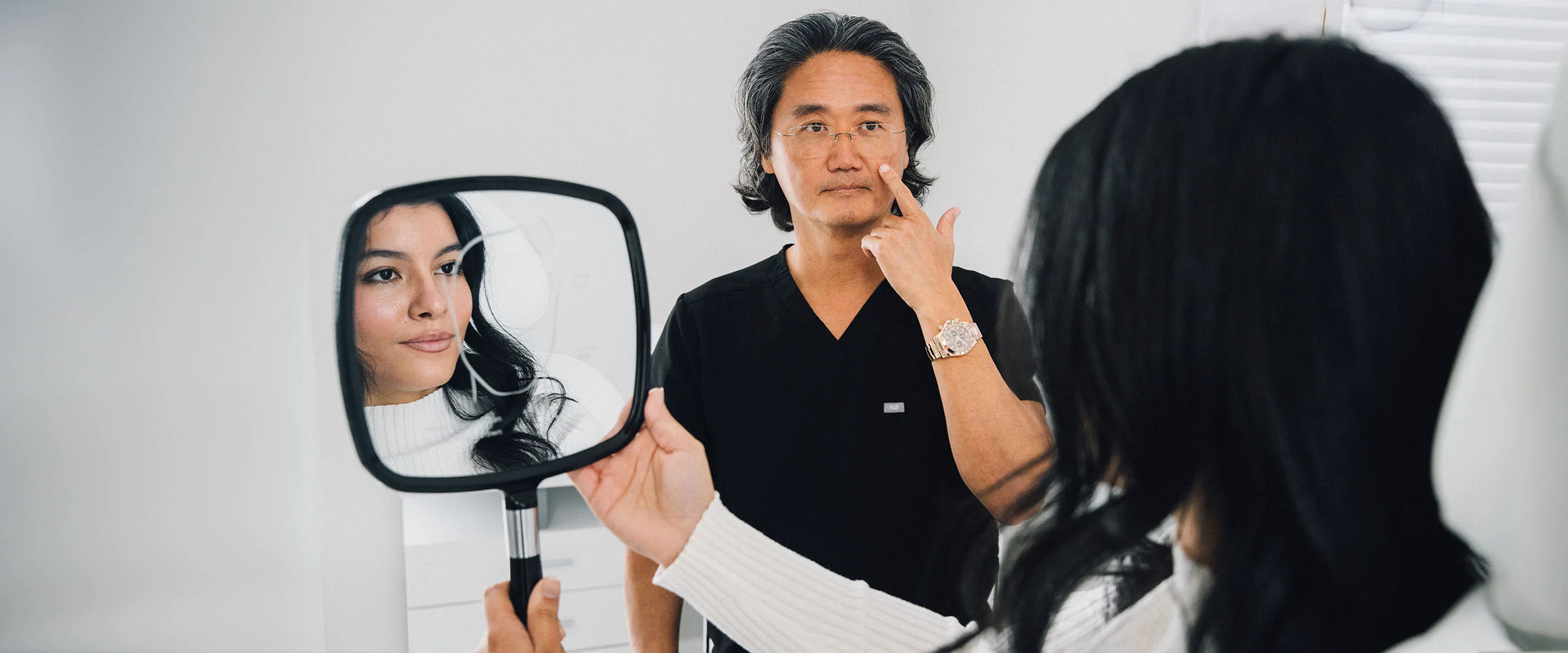 A shot of a woman with long black hair smiling into a handheld mirror as a doctor points at his face, showing her insecure spots. She is wearing a white long shirt, appearing engaged and content during the consultation. - Liquid Facelift in Bryn Mawr, PA