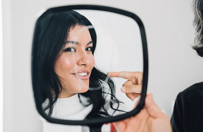 A close-up shot of a woman with long black hair smiling into a handheld mirror as a doctor points at her face. She is wearing a white shirt, appearing engaged and content during the consultation. - Liquid Facelift in Bryn Mawr, PA